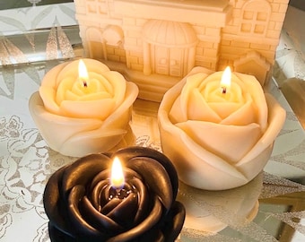 May include: Two cream-colored rose-shaped candles and one black rose-shaped candle are lit, positioned in front of a large, cream-colored, house-shaped candle. The candles feature detailed petal layers and are illuminated with small, bright flames.