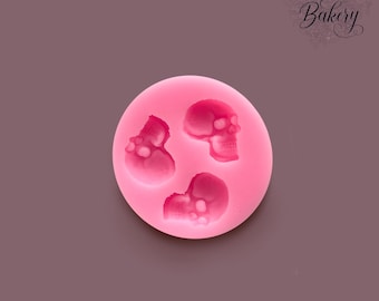 May include: A round, bright pink silicone mold featuring three recessed skull shapes of varying sizes. The mold has a smooth, flexible surface with clearly defined cavities for casting. The text "MY LITTLE Bakery" is visible in the top right corner.