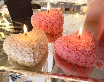 May include: Three heart-shaped candles with a textured floral surface, each featuring a lit wick. One candle is white, while two are shades of pink. The candles are arranged on a reflective surface alongside a dark brown and a white house-shaped object.