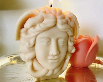 May include: A cream-colored wax candle shaped like a classical human head with detailed wavy hair and a lit wick at the top. Beside it sits a smaller, solid peach-colored candle shaped like a rosebud, both resting on a reflective silver surface.