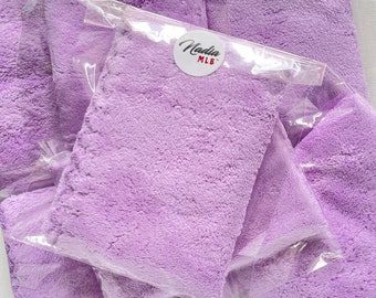Op de afbeelding: Een stapel lavendelkleurige microvezeldoeken in doorzichtige plastic verpakking. De verpakking heeft een ronde sticker met de tekst "Nadia MLB" erop.