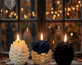 May include: Three pinecone-shaped candles sit on a wooden surface. The left candle is cream, the center is a two-tone cream and dark blue, and the right is dark blue. Each candle has a lit wick, creating a warm glow against a window background.