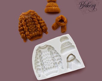 May include: A white silicone mold featuring cavities shaped like a sweater, a beanie, and a scarf, accompanied by three finished amber-colored wax or chocolate figures of the same items. The text My Little Bakery is visible in the upper right corner.