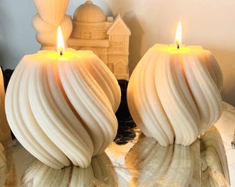 May include: Two off-white, spherical candles with a vertical, spiraling, ribbed texture. Each candle is lit, featuring a single flame at the top center. The wax surface is smooth and matte, reflecting light along the curved, twisted ridges.