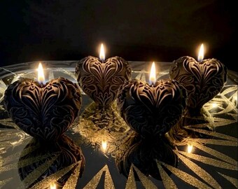 May include: Four black heart-shaped candles with intricate embossed scrollwork patterns. Each candle is lit, producing a bright flame that illuminates the dark surface. The candles are arranged in a group, reflecting light on the glossy base below.