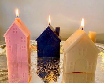 May include: Three house-shaped candles in pink, dark blue, and cream, each with a lit wick. The candles feature molded details including windows and doors, and they are positioned on a reflective surface that shows their glowing reflections.