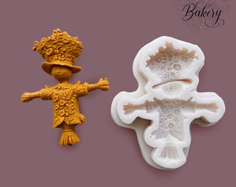 May include: A mustard yellow scarecrow figurine with a floral hat and shirt, alongside a white silicone mold that creates the same scarecrow shape. The scarecrow features a wide-brimmed hat, outstretched arms, and a textured, straw-like base.