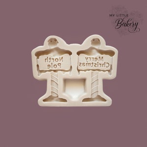 Puede incluir: Un molde de silicona blanco para hornear golosinas con temática navideña. El molde presenta dos figuras que sostienen carteles que dicen "Polo Norte" y "Feliz Navidad".