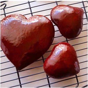 May include: Three glossy, heart-shaped objects in varying sizes, arranged on a black wire rack. The hearts have a deep red color with a shiny, reflective surface, suggesting a glaze or coating. The composition evokes a theme of love or confectionery.