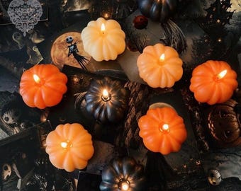 May include: A collection of pumpkin-shaped candles in shades of orange, cream, and black, each featuring a lit wick. The candles are arranged on a dark, patterned surface decorated with illustrations of skeletons, haunted houses, and full moons.