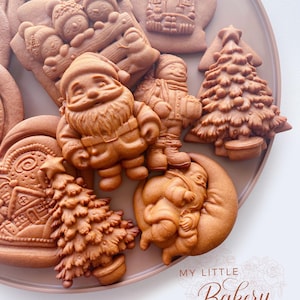 May include: A selection of elaborately decorated gingerbread biscuits in festive shapes, such as Santa Claus, Christmas trees, snowmen, and a house. The biscuits are presented on a brown plate, with the words "MY LITTLE Bakery" in a decorative font.