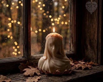 May include: A cream-colored wax candle shaped like a bust of a figure draped in a veil, featuring a small flame at the top. The candle is positioned on a dark wooden surface surrounded by scattered brown autumn leaves near a window pane.