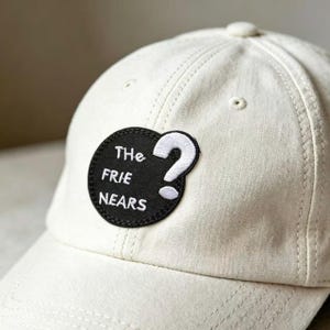 May include: An off-white baseball cap featuring a circular black patch on the front. The patch is embroidered with white text that reads "THe FRIE NEARS" and includes a large white question mark symbol to the right of the lettering.