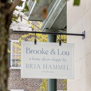 May include: A rectangular, light-coloured hanging sign suspended from a black metal rod. The sign features dark blue text that reads Brooke & Lou, a home décor shoppe by BRIA HAMMEL INTERIORS, framed by a thin white border on a light background.