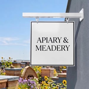 May include: A square white hanging sign with a black border and the black text APIARY & MEADERY. The sign is suspended from a white metal bracket attached to a gray wall, positioned above a garden area with wooden beehives and yellow flowers.