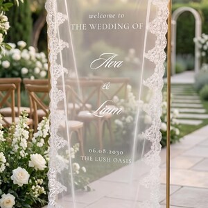 Pu&ograve; includere: Un alto telaio rettangolare in metallo dorato che sostiene un pannello in tessuto bianco trasparente, rifinito con un intricato bordo in pizzo bianco. Il testo stampato in bianco recita: welcome to THE WEDDING OF Ava & Liam 06.08.2030 THE LUSH OASIS.