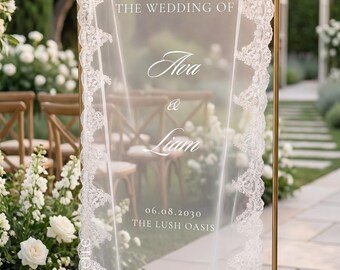 Può includere: Un alto telaio rettangolare in metallo dorato che sostiene un pannello in tessuto bianco trasparente, rifinito con un intricato bordo in pizzo bianco. Il testo stampato in bianco recita: welcome to THE WEDDING OF Ava & Liam 06.08.2030 THE LUSH OASIS.