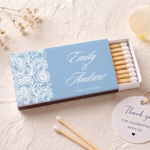 K&ouml;nnte beinhalten: Eine hellblaue Streichholzschachtel mit einem wei&szlig;en floralen Spitzenmuster auf der linken Seite und dem Text Emily & Andrew May 25th 2030 in wei&szlig;er Schreibschrift. Die Schachtel ist leicht ge&ouml;ffnet und zeigt Streichh&ouml;lzer mit wei&szlig;en K&ouml;pfen.