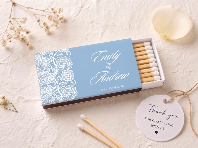 K&ouml;nnte beinhalten: Eine hellblaue Streichholzschachtel mit einem wei&szlig;en floralen Spitzenmuster auf der linken Seite und dem Text Emily & Andrew May 25th 2030 in wei&szlig;er Schreibschrift. Die Schachtel ist leicht ge&ouml;ffnet und zeigt Streichh&ouml;lzer mit wei&szlig;en K&ouml;pfen.