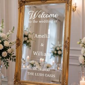 May include: A rectangular mirror with an ornate gold frame, featuring white text that reads: Welcome to the wedding of Amelia & William 06.08.2030 THE LUSH OASIS. The mirror is mounted on a wooden easel, reflecting a room with white flowers and candles.