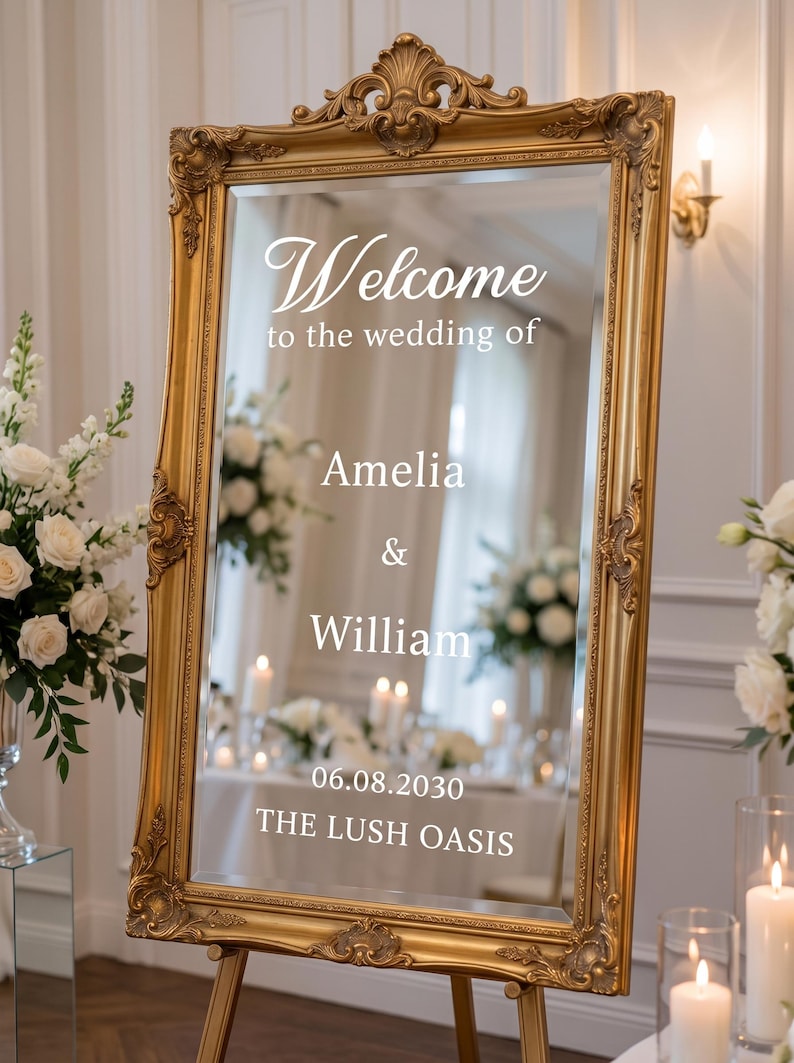 May include: A rectangular mirror with an ornate gold frame, featuring white text that reads: Welcome to the wedding of Amelia & William 06.08.2030 THE LUSH OASIS. The mirror is mounted on a wooden easel, reflecting a room with white flowers and candles.