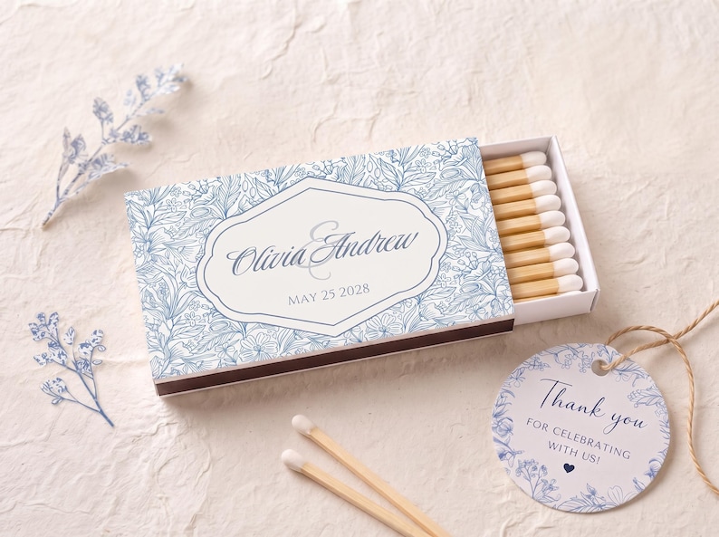 K&ouml;nnte beinhalten: Eine rechteckige Streichholzschachtel mit blau-wei&szlig;em Blumenmuster und dem Text Olivia & Andrew sowie MAY 25 2028 in einem zentralen Rahmen. Die Schachtel enth&auml;lt Streichh&ouml;lzer mit wei&szlig;en K&ouml;pfen. Ein rundes Etikett tr&auml;gt den Text Thank you FOR CELEBRATING WITH US!