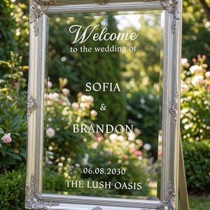May include: A rectangular mirror with an ornate silver frame featuring white text that reads: Welcome to the wedding of SOFIA & BRANDON 06.08.2030 THE LUSH OASIS. The mirror is mounted on a stand and reflects a garden setting with green foliage.