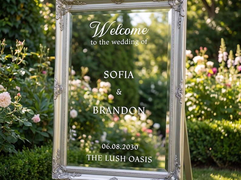 May include: A rectangular mirror with an ornate silver frame featuring white text that reads: Welcome to the wedding of SOFIA & BRANDON 06.08.2030 THE LUSH OASIS. The mirror is mounted on a stand and reflects a garden setting with green foliage.