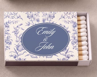 Könnte beinhalten: Eine rechteckige Streichholzschachtel mit cremefarbenem Hintergrund und blauem Blumenmuster. Ein zentrales blaues Oval enthält den weißen Text Emily & John. Die Schachtel ist leicht geöffnet und zeigt eine Reihe von Streichhölzern mit weißen Köpfen.