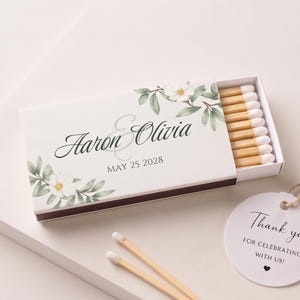 K&ouml;nnte beinhalten: Eine rechteckige Streichholzschachtel mit wei&szlig;em Deckel, Blumenmotiv, den Namen Aaron und Olivia sowie dem Datum 25. Mai 2028. Die Schachtel ist leicht ge&ouml;ffnet und zeigt Streichh&ouml;lzer mit wei&szlig;en K&ouml;pfen. Ein rundes Etikett tr&auml;gt die Aufschrift Thank you for celebrating with us.