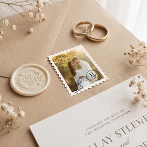 K&ouml;nnte beinhalten: Ein beigefarbener Umschlag mit einer personalisierten Briefmarke, die ein Foto eines Paares zeigt, einem cremefarbenen Wachssiegel mit gepr&auml;gtem Blumenmonogramm und zwei goldenen Eheringen. Eine wei&szlig;e Hochzeitseinladungskarte ist teilweise sichtbar.