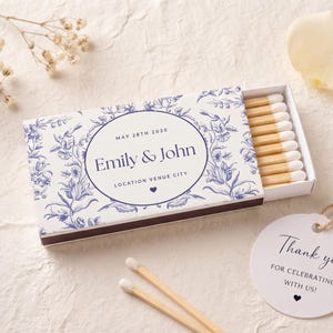 K&ouml;nnte beinhalten: Eine rechteckige Streichholzschachtel mit blau-wei&szlig;em Blumenmuster und dem Text "Emily & John", "MAY 28TH 2030" sowie "LOCATION VENUE CITY". Die Schachtel enth&auml;lt Holzstreichh&ouml;lzer mit wei&szlig;en K&ouml;pfen. Ein rundes wei&szlig;es Etikett tr&auml;gt die Aufschrift "Thank you FOR CELEBRATING WITH US!"