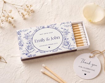 Könnte beinhalten: Eine rechteckige Streichholzschachtel mit blau-weißem Blumenmuster und dem Text "Emily & John", "MAY 28TH 2030" sowie "LOCATION VENUE CITY". Die Schachtel enthält Holzstreichhölzer mit weißen Köpfen. Ein rundes weißes Etikett trägt die Aufschrift "Thank you FOR CELEBRATING WITH US!"