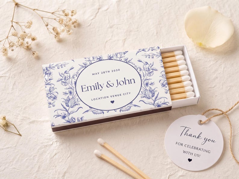 K&ouml;nnte beinhalten: Eine rechteckige Streichholzschachtel mit blau-wei&szlig;em Blumenmuster und dem Text "Emily & John", "MAY 28TH 2030" sowie "LOCATION VENUE CITY". Die Schachtel enth&auml;lt Holzstreichh&ouml;lzer mit wei&szlig;en K&ouml;pfen. Ein rundes wei&szlig;es Etikett tr&auml;gt die Aufschrift "Thank you FOR CELEBRATING WITH US!"