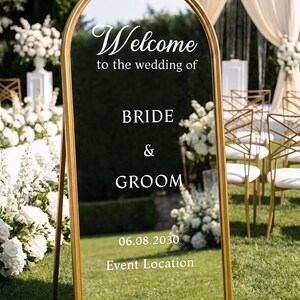 May include: An arched floor mirror with a gold frame featuring white text that reads Welcome to the wedding of BRIDE & GROOM 06.08.2030 Event Location. The mirror reflects a grassy outdoor setting with white floral arrangements and chairs.