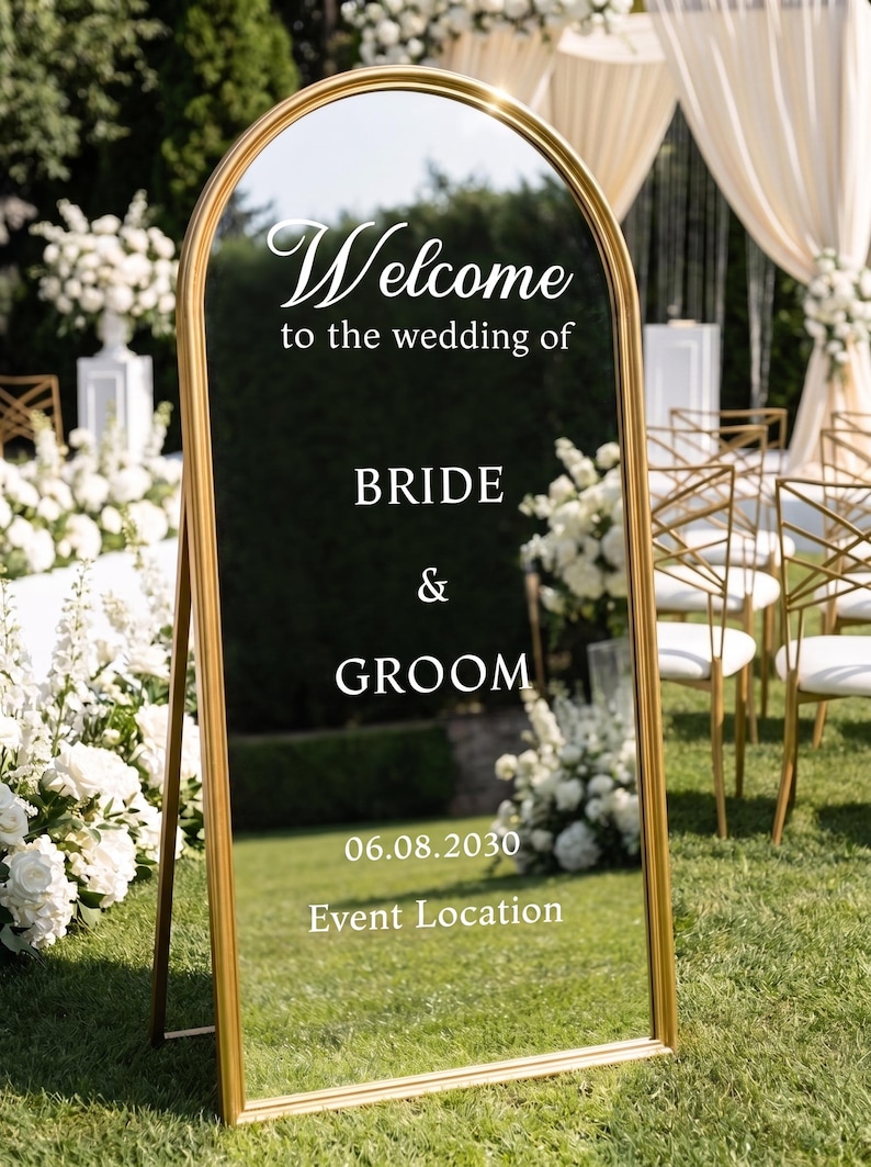 May include: An arched floor mirror with a gold frame featuring white text that reads Welcome to the wedding of BRIDE & GROOM 06.08.2030 Event Location. The mirror reflects a grassy outdoor setting with white floral arrangements and chairs.