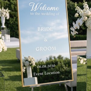 May include: A rectangular mirror wedding sign with a gold frame, mounted on a silver easel. The white text reads: Welcome to the wedding of BRIDE & GROOM 06.08.2030 Event Location. The mirror reflects the surrounding outdoor green lawn and sky.