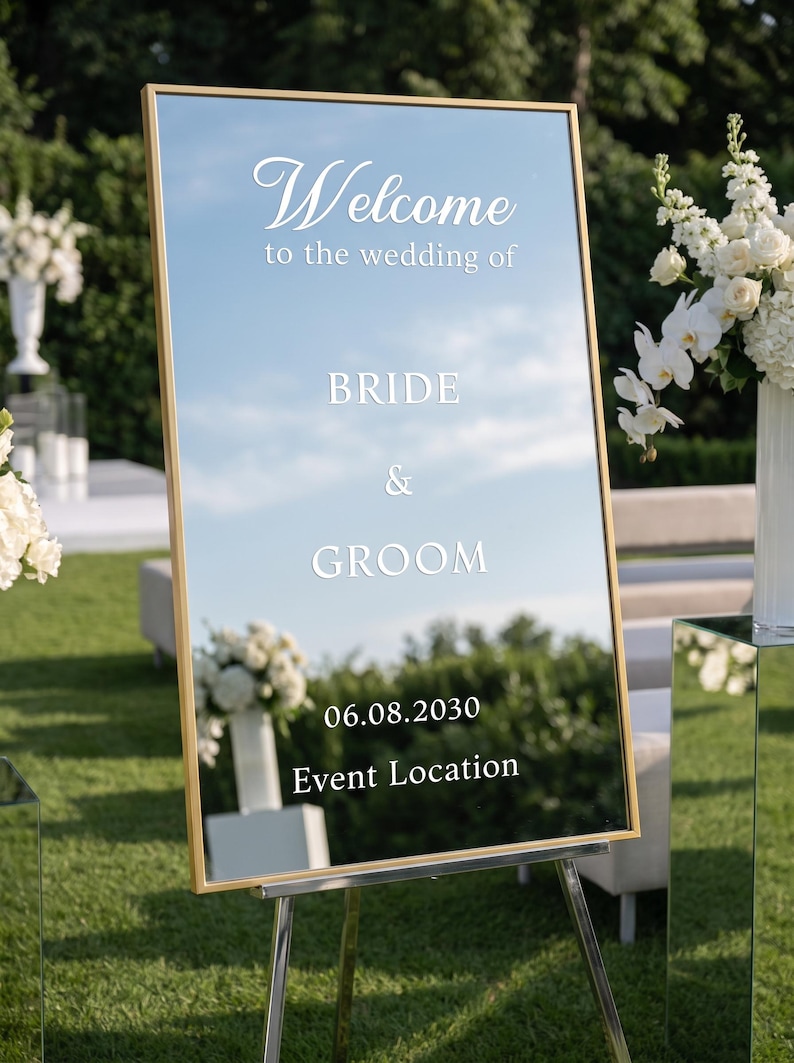 May include: A rectangular mirror wedding sign with a gold frame, mounted on a silver easel. The white text reads: Welcome to the wedding of BRIDE & GROOM 06.08.2030 Event Location. The mirror reflects the surrounding outdoor green lawn and sky.