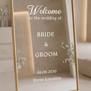 May include: A rectangular mirror sign with a thin gold frame featuring white text that reads Welcome to the wedding of BRIDE & GROOM 06.08.2030 Event Location. The reflective surface displays white floral accents on the sides.