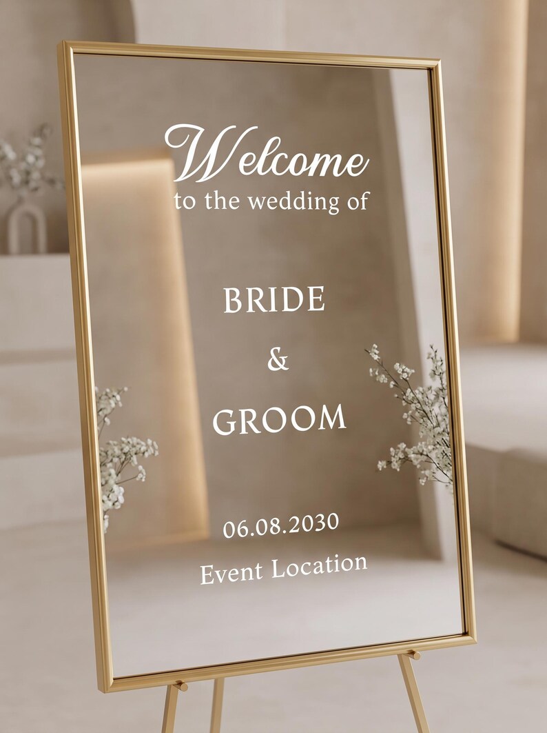 May include: A rectangular mirror sign with a thin gold frame featuring white text that reads Welcome to the wedding of BRIDE & GROOM 06.08.2030 Event Location. The reflective surface displays white floral accents on the sides.