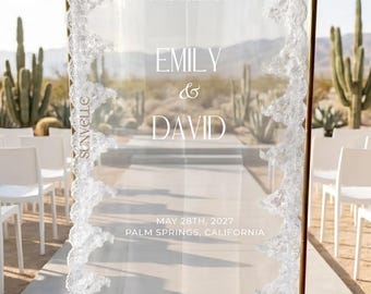 Può includere: Un alto cartello di benvenuto per matrimoni di forma rettangolare, con un pannello in tessuto bianco trasparente drappeggiato all'interno di una cornice in metallo dorato. Il tessuto è bordato da un intricato pizzo bianco. Il testo recita: WELCOME TO THE WEDDING OF EMILY & DAVID MAY 28TH, 2027 PALM SPRINGS, CALIFORNIA.