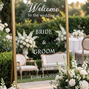 May include: A tall rectangular mirror with a gold frame featuring white text that reads Welcome to the wedding of BRIDE & GROOM 06.08.2030 Event Location. The mirror reflects a garden setting with white flowers and outdoor furniture.
