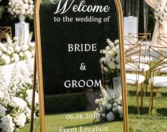 Può includere: Uno specchio da terra ad arco con cornice dorata che presenta una scritta bianca che recita Welcome to the wedding of BRIDE & GROOM 06.08.2030 Event Location. Lo specchio riflette un ambiente esterno erboso con composizioni floreali bianche e sedie.