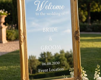 May include: A rectangular mirror with an ornate gold frame featuring white text that reads Welcome to the wedding of BRIDE & GROOM 06.08.2030 Event Location. The mirror is mounted on a stand and reflects the surrounding outdoor greenery.