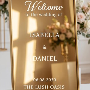 May include: A rectangular gold-toned mirror sign with a thin gold frame. The white text reads: Welcome to the wedding of ISABELLA & DANIEL 06.08.2030 THE LUSH OASIS. The reflective surface shows light reflections and a blurred floral arrangement.