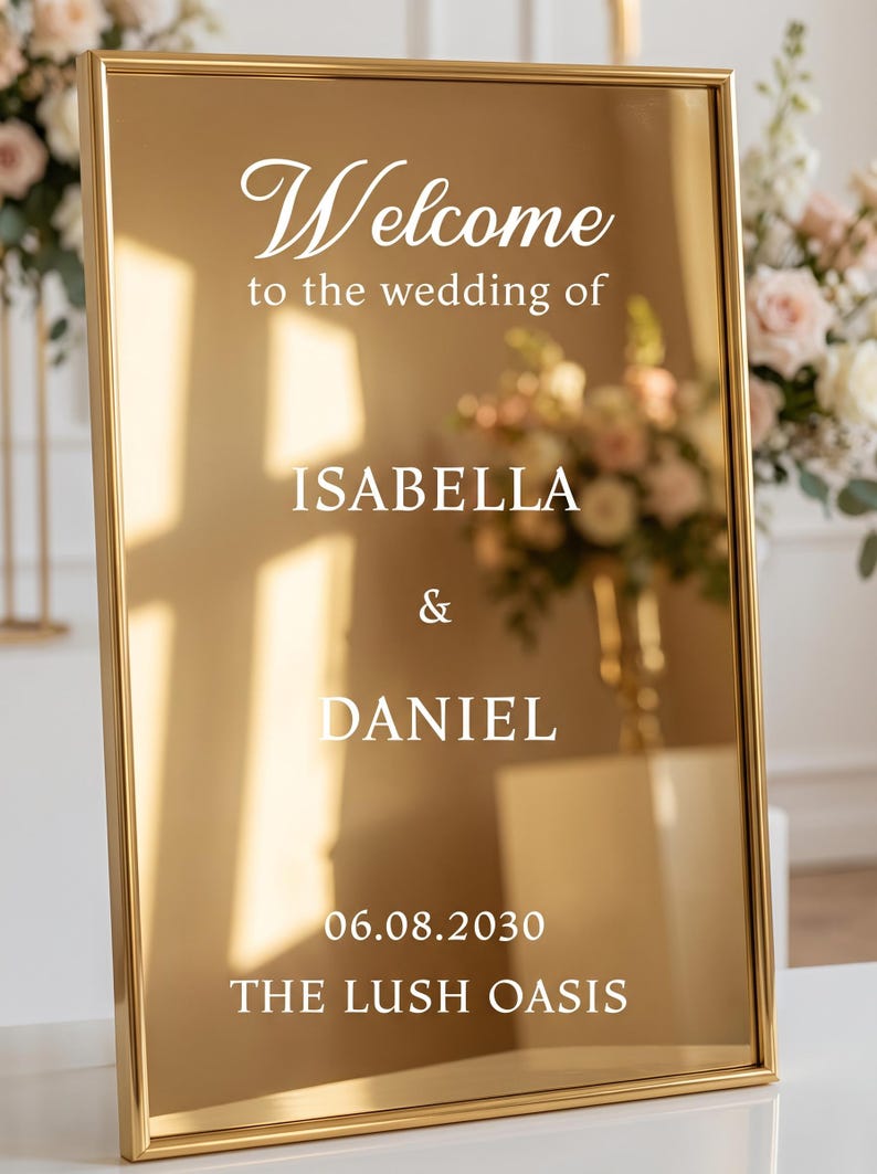 May include: A rectangular gold-toned mirror sign with a thin gold frame. The white text reads: Welcome to the wedding of ISABELLA & DANIEL 06.08.2030 THE LUSH OASIS. The reflective surface shows light reflections and a blurred floral arrangement.