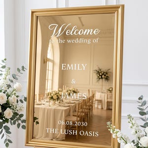 May include: A rectangular gold-framed mirror sign with white text that reads: Welcome to the wedding of EMILY & JAMES 06.08.2030 THE LUSH OASIS. The mirror reflects a room with tables, chairs, and floral arrangements, standing on a gold easel.