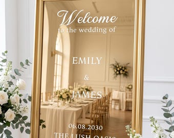 Può includere: Uno specchio rettangolare con cornice dorata e scritta bianca: Welcome to the wedding of EMILY & JAMES 06.08.2030 THE LUSH OASIS. Lo specchio riflette una stanza con tavoli, sedie e fiori, ed è posizionato su un cavalletto dorato.