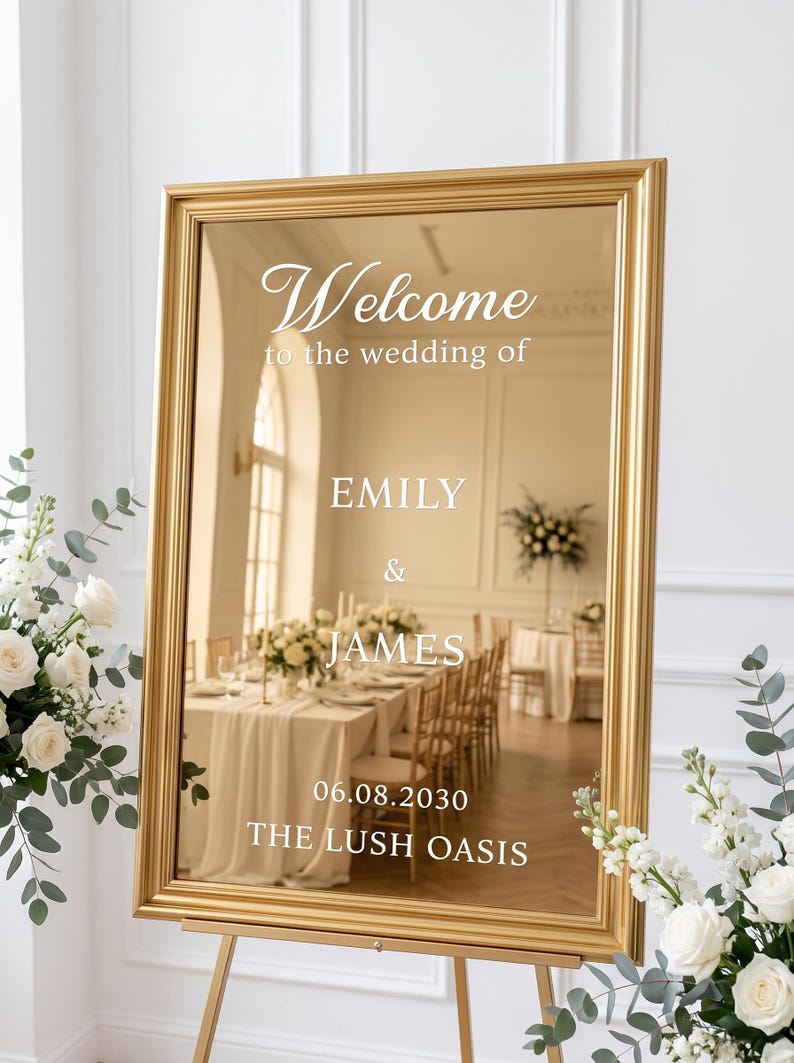 May include: A rectangular gold-framed mirror sign with white text that reads: Welcome to the wedding of EMILY & JAMES 06.08.2030 THE LUSH OASIS. The mirror reflects a room with tables, chairs, and floral arrangements, standing on a gold easel.