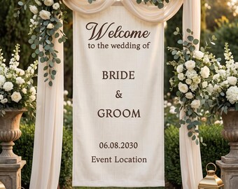 May include: A wedding welcome sign hanging from a wooden arch draped with cream fabric and decorated with white roses and eucalyptus. The banner features dark brown text reading Welcome to the wedding of BRIDE & GROOM 06.08.2030 Event Location.